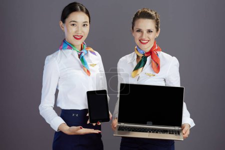 Photo for Smiling stylish air hostess women in blue skirt, white shirt and scarf showing tablet PC blank screen and and laptop blank screen isolated on grey. - Royalty Free Image