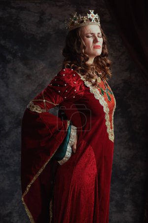 Photo for Tired medieval queen in red dress with crown having backache on dark gray background. - Royalty Free Image