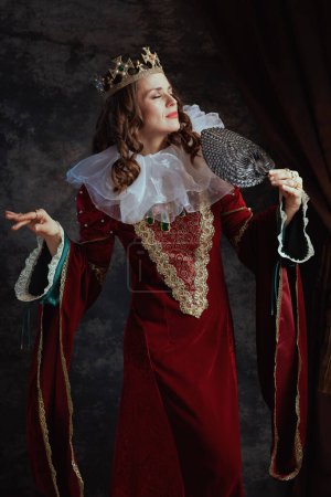 Photo for Medieval queen in red dress with fan, white collar and crown on dark gray background. - Royalty Free Image