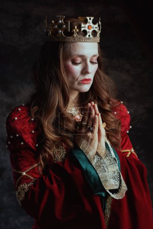 Téléchargez les photos : Reine médiévale en robe rouge avec couronne priant sur fond gris foncé. - en image libre de droit