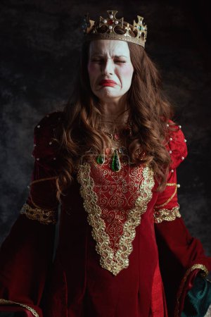 Photo for Sad medieval queen in red dress with white makeup and crown on dark gray background. - Royalty Free Image