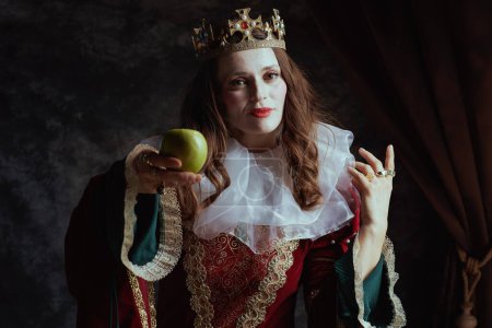 Téléchargez les photos : Reine médiévale en robe rouge avec pomme verte, col blanc et couronne sur fond gris foncé. - en image libre de droit