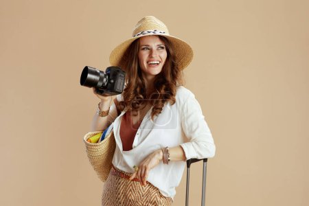 Téléchargez les photos : Vacances à la plage. heureuse femme élégante en chemisier blanc et short isolé sur fond beige avec sac de paille, sac trolley, appareil photo et chapeau de paille. - en image libre de droit