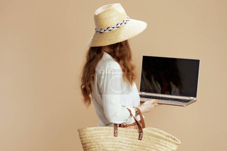 Téléchargez les photos : Vacances à la plage. heureuse femme élégante en chemisier blanc et short isolé sur fond beige avec sac de paille et chapeau de paille montrant écran blanc ordinateur portable. - en image libre de droit