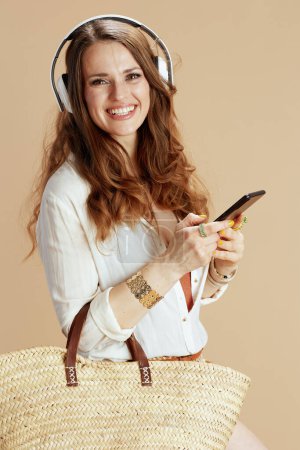 Beach vacation. happy elegant 40 years old housewife in white blouse and shorts isolated on beige with straw bag listening to the music with headphones and using smartphone.