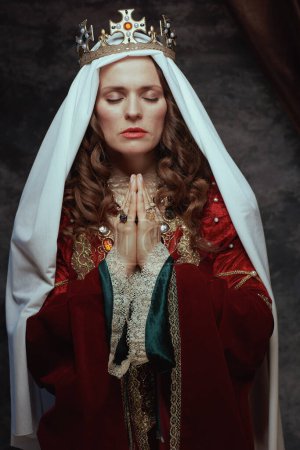 Photo for Medieval queen in red dress with veil and crown praying on dark gray background. - Royalty Free Image