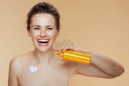 Photo for Smiling modern woman with cosmetic cream jar and heart shaped cream on chest isolated on beige background. - Royalty Free Image