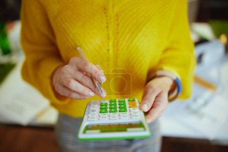Primer plano de la mujer propietaria de una pequeña empresa en suéter amarillo con calculadora en la oficina verde.