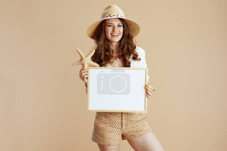Photo for Beach vacation. happy stylish 40 years old housewife in white blouse and shorts on beige background with sea star and straw hat showing blank board. - Royalty Free Image
