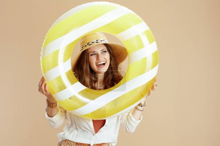 Photo for Beach vacation. happy elegant middle aged woman in white blouse and shorts isolated on beige with inflatable circle and summer hat. - Royalty Free Image