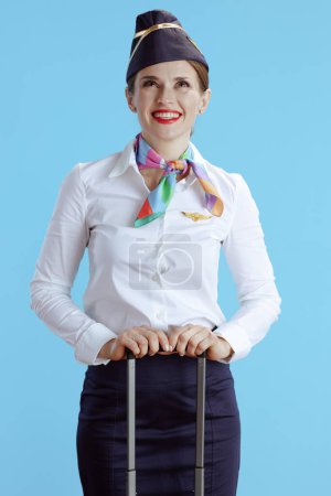 Foto de Elegante asistente de vuelo femenino sonriente aislado sobre fondo azul en uniforme con bolsa de viaje mirando hacia arriba en el espacio de copia. - Imagen libre de derechos