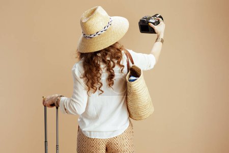 Téléchargez les photos : Vacances à la plage. Vu de derrière femme en chemisier blanc et short sur fond beige avec sac de paille, sac trolley, appareil photo et chapeau de paille. - en image libre de droit