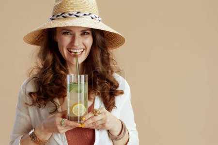 Photo for Beach vacation. smiling elegant 40 years old woman in white blouse and shorts against beige background with cocktail and straw hat. - Royalty Free Image