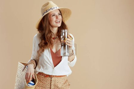 Téléchargez les photos : Vacances à la plage. heureux élégant femme en chemisier blanc et short isolé sur beige avec bouteille d'eau, sac de paille et chapeau de paille. - en image libre de droit