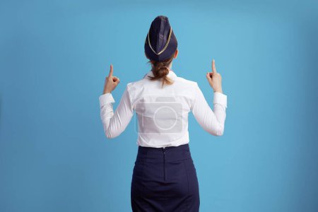 Photo for Seen from behind modern female air hostess against blue background in uniform pointing up. - Royalty Free Image