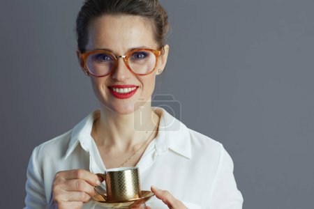 Foto de Sonriente mujer de negocios de moda de 40 años en blusa blanca con gafas y taza de café aislada sobre fondo gris. - Imagen libre de derechos