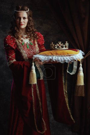 Photo for Medieval queen in red dress with crown on pillow on dark gray background. - Royalty Free Image
