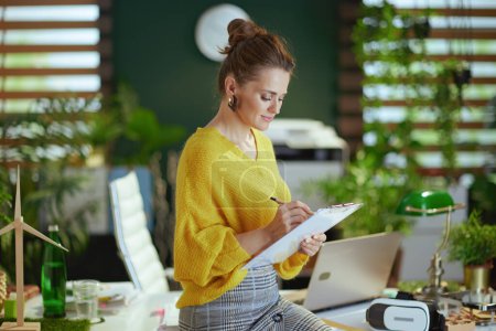 Foto de Mujer propietaria de la pequeña empresa de moda en suéter amarillo con portapapeles en la moderna oficina verde. - Imagen libre de derechos