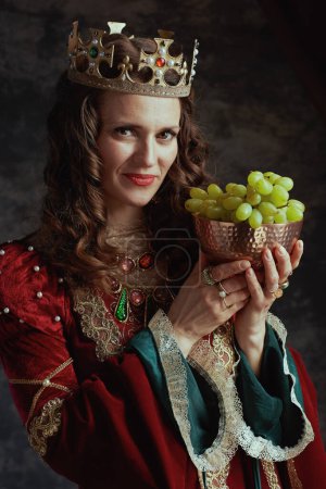 Photo for Happy medieval queen in red dress with plate of grapes and crown on dark gray background. - Royalty Free Image