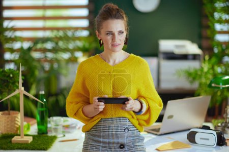 Foto de Elegante mujer de mediana edad propietaria de una pequeña empresa en suéter amarillo en la moderna oficina verde con teléfono inteligente. - Imagen libre de derechos