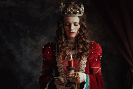 Photo for Medieval queen in red dress with candle and crown on dark gray background. - Royalty Free Image