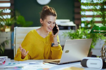 Foto de Sonriendo 40 años de edad propietaria de una pequeña empresa mujer en suéter amarillo con portátil hablando en un teléfono inteligente y trabajando con documentos en la moderna oficina verde. - Imagen libre de derechos