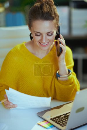 Photo for Happy trendy middle aged small business owner woman in yellow sweater with laptop talking on a smartphone in the modern green office. - Royalty Free Image