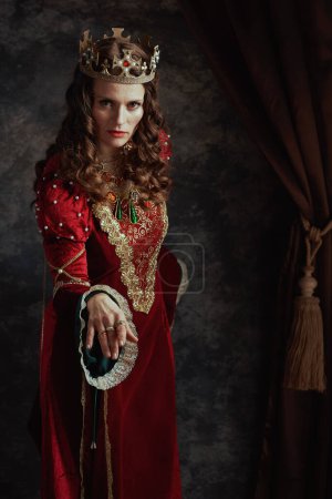 Photo for Medieval queen in red dress with crown making reverence on dark gray background. - Royalty Free Image