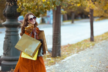 Foto de Hola noviembre. mujer elegante sonriente en gabardina marrón con bolsas de compras y hojas amarillas de otoño en la ciudad. - Imagen libre de derechos