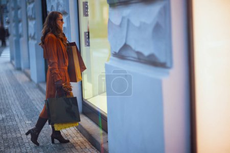 Foto de Hola otoño. Retrato de larga duración de una mujer moderna de 40 años en gabardina naranja con bolsas de compras y hojas amarillas de otoño cerca de la tienda en la ciudad. - Imagen libre de derechos