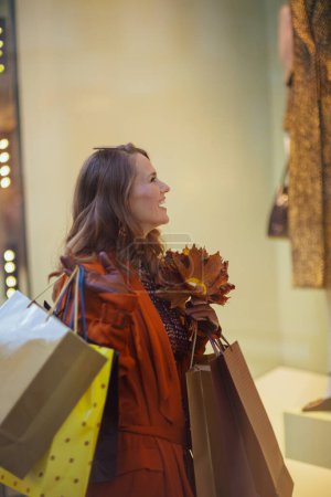 Foto de Hola septiembre. sonriente mujer moderna de mediana edad en gabardina marrón con bolsas de compras y hojas amarillas de otoño cerca de la tienda en la ciudad. - Imagen libre de derechos