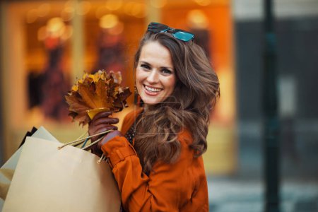 Foto de Hola octubre. hembra sonriente de moda en gabardina marrón con bolsas de compras y hojas amarillas de otoño cerca de la tienda en la ciudad. - Imagen libre de derechos