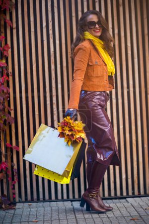 Foto de Hola septiembre. Retrato completo de mujer elegante sonriente en gabardina marrón con bolsas de compras y hojas amarillas de otoño en la ciudad. - Imagen libre de derechos
