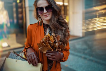 Téléchargez les photos : Bonjour octobre. jeune femme en trench-coat marron avec des sacs à provisions et des feuilles jaune automne près du magasin dans la ville. - en image libre de droit