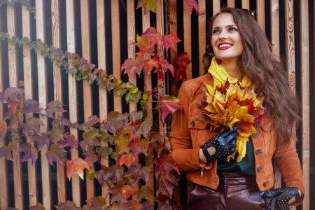 Foto de Hola noviembre. mujer de 40 años de moda relajada en gabardina naranja con hojas amarillas otoñales en la ciudad. - Imagen libre de derechos