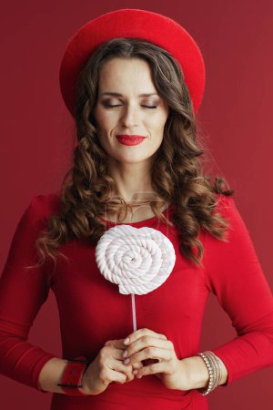 Photo for Happy Valentine. relaxed stylish 40 years old woman in red dress and beret with candy on stick and long wavy hair isolated on red. - Royalty Free Image