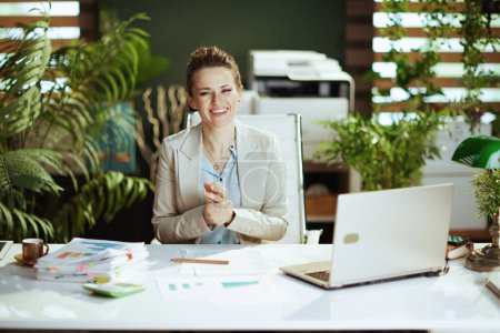 Photo for Portrait of smiling modern middle aged bookkeeper woman in a light business suit in modern green office with laptop. - Royalty Free Image