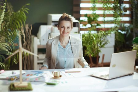 Téléchargez les photos : Portrait de femme comptable moderne heureuse dans un costume d'affaires léger dans un bureau vert moderne avec ordinateur portable. - en image libre de droit