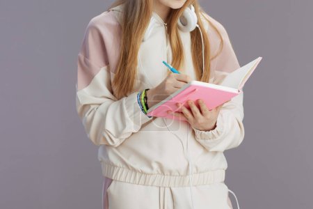 Foto de Primer plano de la chica adolescente en chándal beige con libros de trabajo y auriculares escrito aislado sobre fondo gris. - Imagen libre de derechos