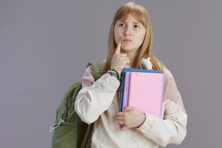 Foto de Chica adolescente moderna pensativa en chándal beige con mochila, libros de trabajo y auriculares aislados en gris. - Imagen libre de derechos