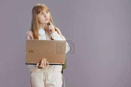 Photo for Pensive modern school girl in beige tracksuit with headphones and laptop isolated on grey. - Royalty Free Image