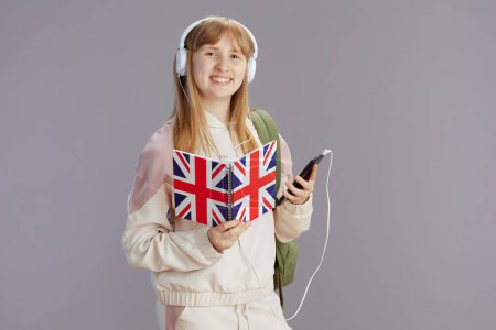 Foto de Niña sonriente de moda en chándal beige con mochila, libro de trabajo de la bandera inglesa y auriculares con teléfono inteligente aislado en gris. - Imagen libre de derechos