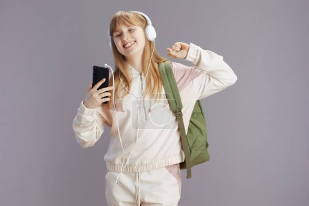 Foto de Pupila de moda feliz en chándal beige con mochila escuchando la música con auriculares y utilizando la aplicación de teléfono inteligente contra gris. - Imagen libre de derechos