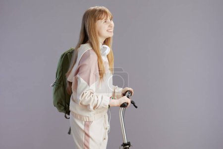Foto de Sonriente mujer joven de moda en chándal beige con mochila, auriculares y scooter aislados sobre fondo gris. - Imagen libre de derechos