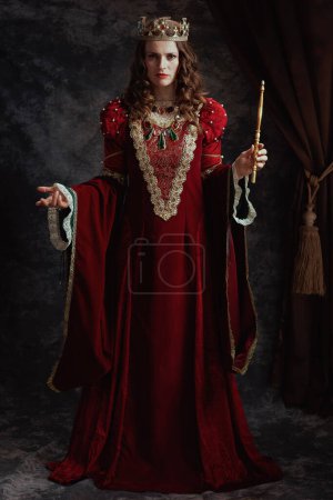Full length portrait of medieval queen in red dress with wand and crown on dark gray background.