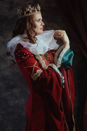 Foto de Reina medieval pensativa en vestido rojo con cuello blanco y corona sobre fondo gris oscuro. - Imagen libre de derechos