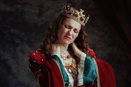 Photo for Stressed medieval queen in red dress with crown having neck pain on dark gray background. - Royalty Free Image