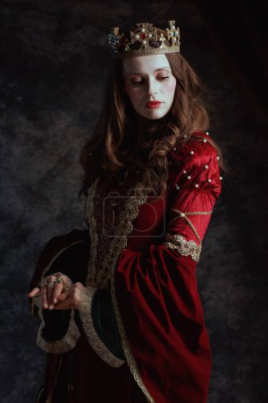 Téléchargez les photos : Reine médiévale en robe rouge avec maquillage blanc et couronne sur fond gris foncé. - en image libre de droit