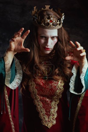 Photo for Scary medieval queen in red dress with white makeup and crown on dark gray background. - Royalty Free Image