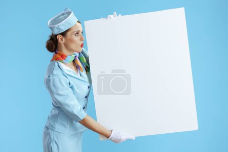 Photo for Surprised stylish female stewardess against blue background in blue uniform showing blank board. - Royalty Free Image